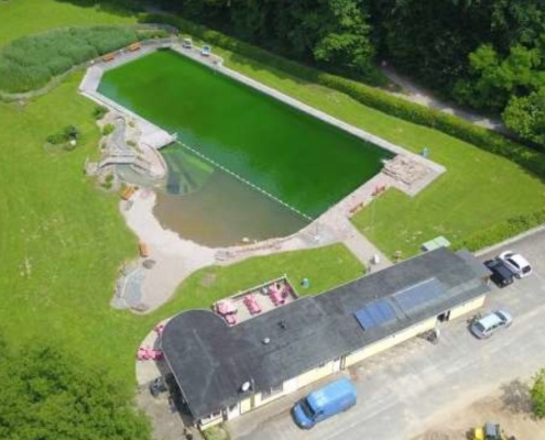 Drohnenbild des Naturbadesees im Hintergrund und einem zweistöckigen Gebäude im Vordergrund.